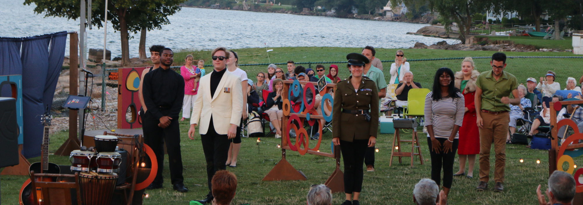 Actors standing facing audience in an outdoor venue with a lake in the background
