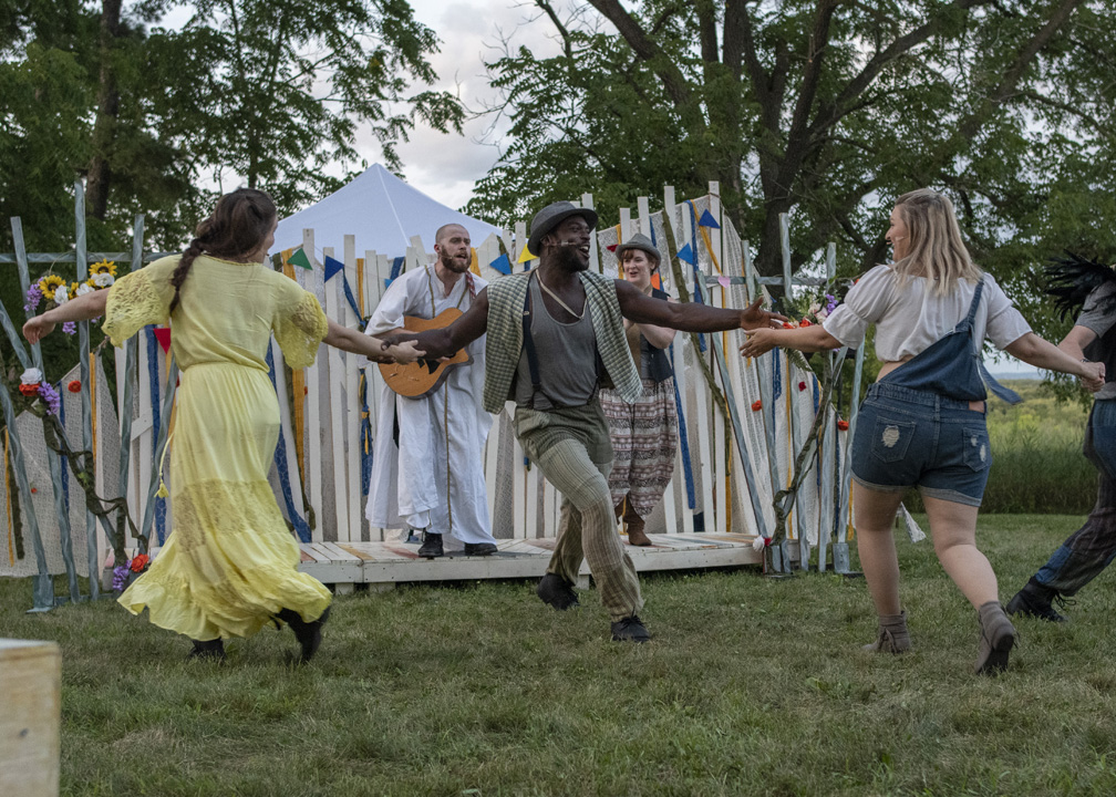 three actors dancing, two white women and one Black man and one white man playing a guitar in the background during a production of As You Like It