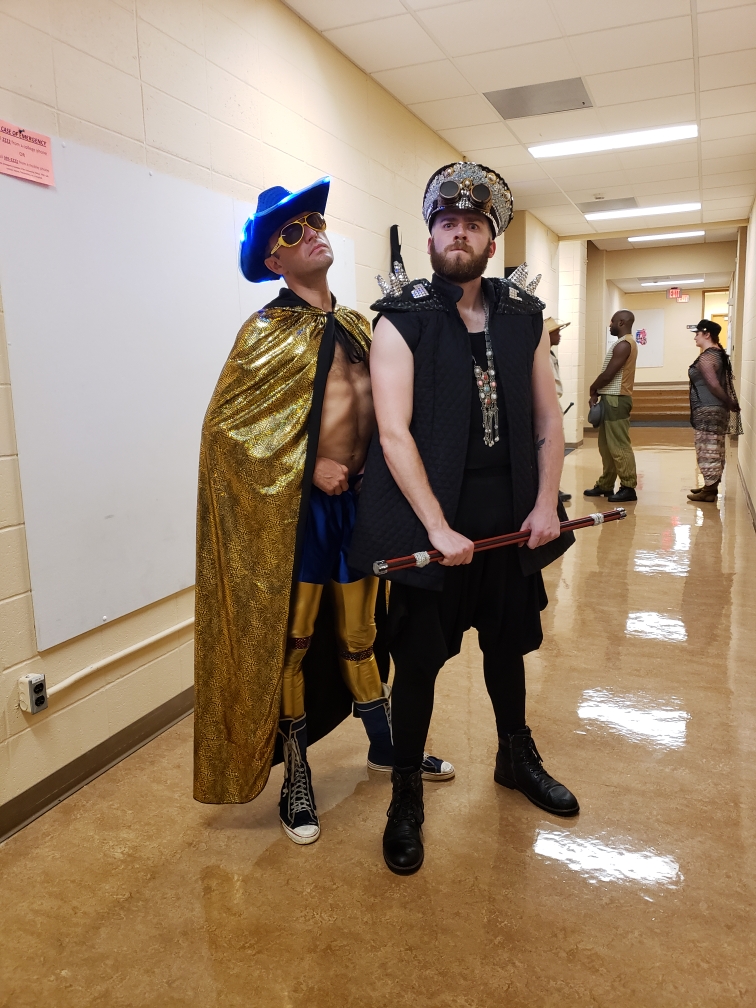 Two white males standing next to each other in a hallway. One is wearing a gold cape with a blue cowboy hat. The other is wearing a black outfit.