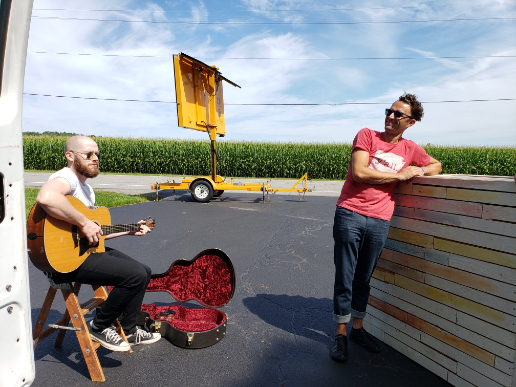 One white male sitting playing guitar and one white male standing leaning up against a platform sitting outside with a cornfield behind them