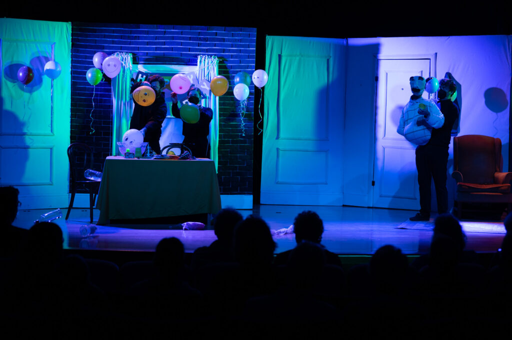 Balloons with faces on them torment a puppet with a puppeteer at a school auditorium